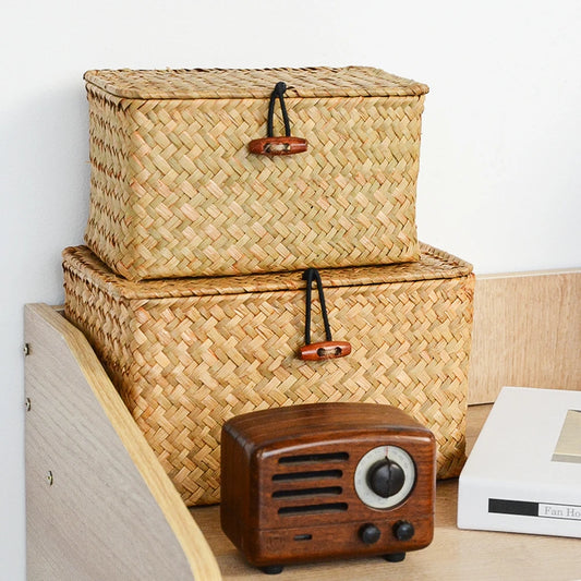 Woven storage baskets with wooden handles on a surface next to a small wooden radio. Handmade seagrass rattan storage box with lid, rectangular woven organizer basket for makeup, jewelry, office, and boho home decor