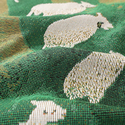 Close-up of fabric with white animal pattern on green background Grassland sheep tassel blanket, woven scenic throw for sofa, bed, or outdoor picnic, boho-inspired home decor blanket.