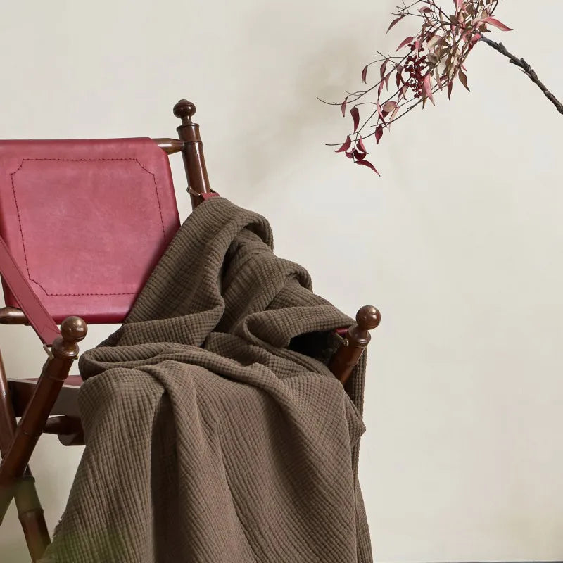 Brown blanket draped over a wooden chair with pink cushion against a neutral wall. Maillard cotton gauze blanket with tassels, four-layer muslin throw for sofa, bed, and boho home decor