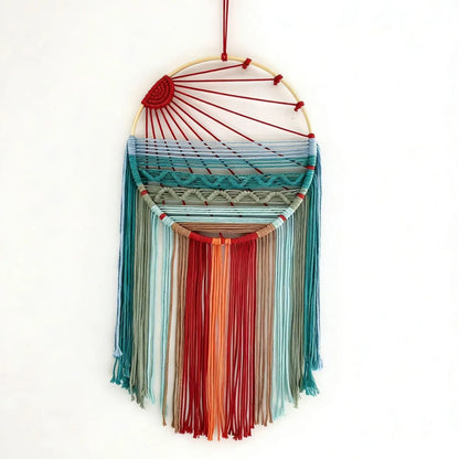A bohemian-style dreamcatcher wall hanging suspended from a thin black string, featuring a natural wood hoop, red wrapped cords, and a multi-colored woven web with long fringe tassels in shades of red, orange, teal, and blue against a plain white background.