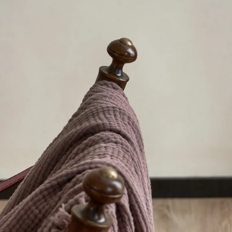 Close-up of a wooden coat rack with a brown towel draped over it against a neutral background. Maillard cotton gauze blanket with tassels, four-layer muslin throw for sofa, bed, and boho home decor