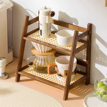 Wooden shelf with kitchen items including a coffee maker, cups, and a pitcher on a light wooden floor. 2-tier bamboo countertop organizer shelf in walnut and blonde finish, multipurpose coffee cup holder and storage rack for kitchen, bathroom, or vanity