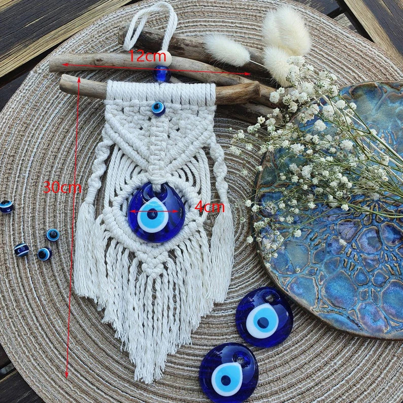 Macrame wall hanging with blue evil eye beads on a textured surface with dried flowers and branches.