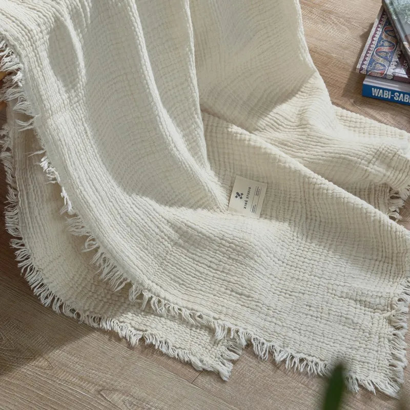 White textured blanket with fringes on a wooden floor Maillard cotton gauze blanket with tassels, four-layer muslin throw for sofa, bed, and boho home decor