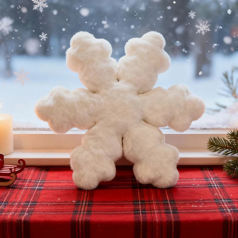 A fluffy white snowflake-shaped plush toy or decorative pillow sits on a red plaid table runner in front of a window with snow visible outside.
