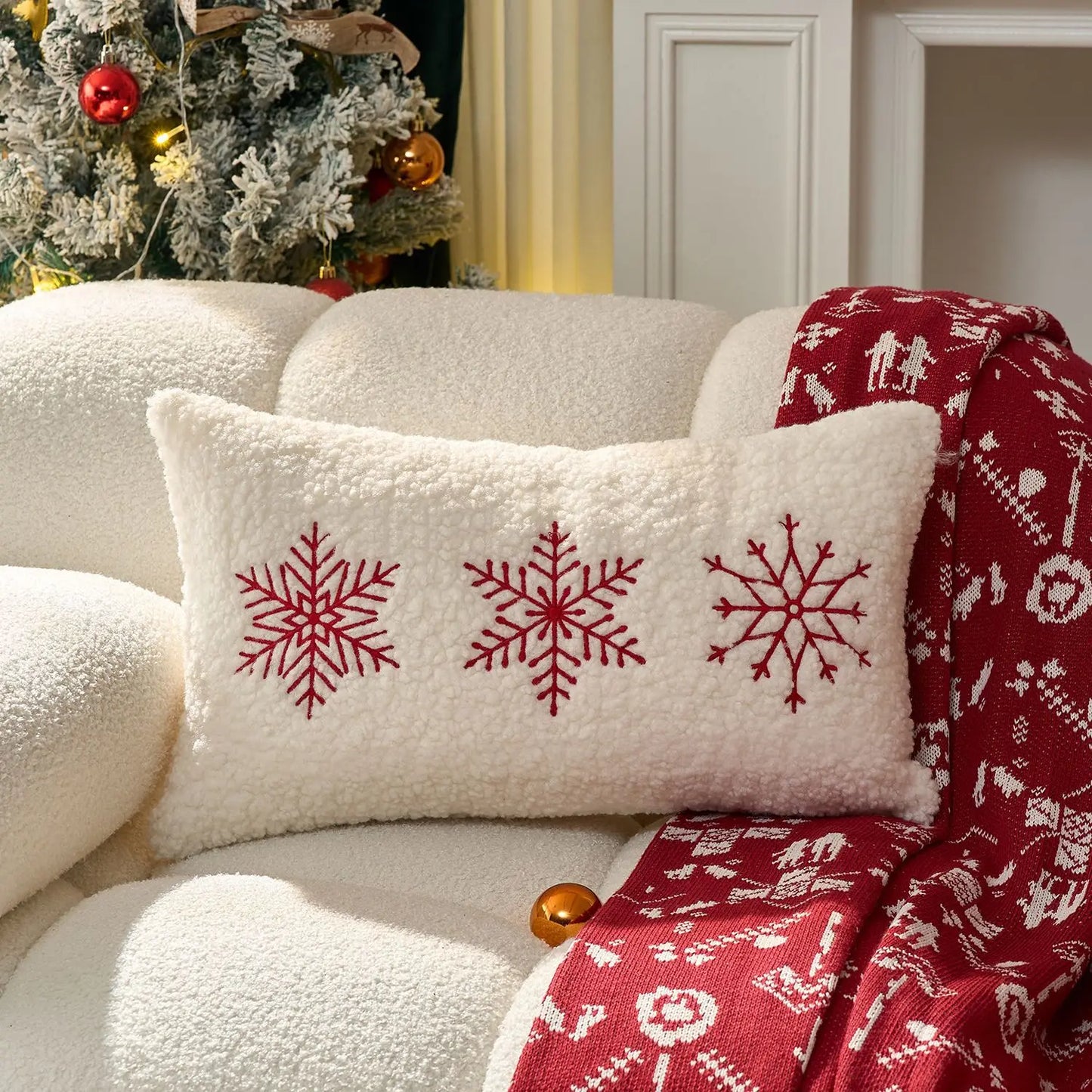 Decorative pillow with red snowflakes on a white couch with a red blanket and Christmas tree in the background.