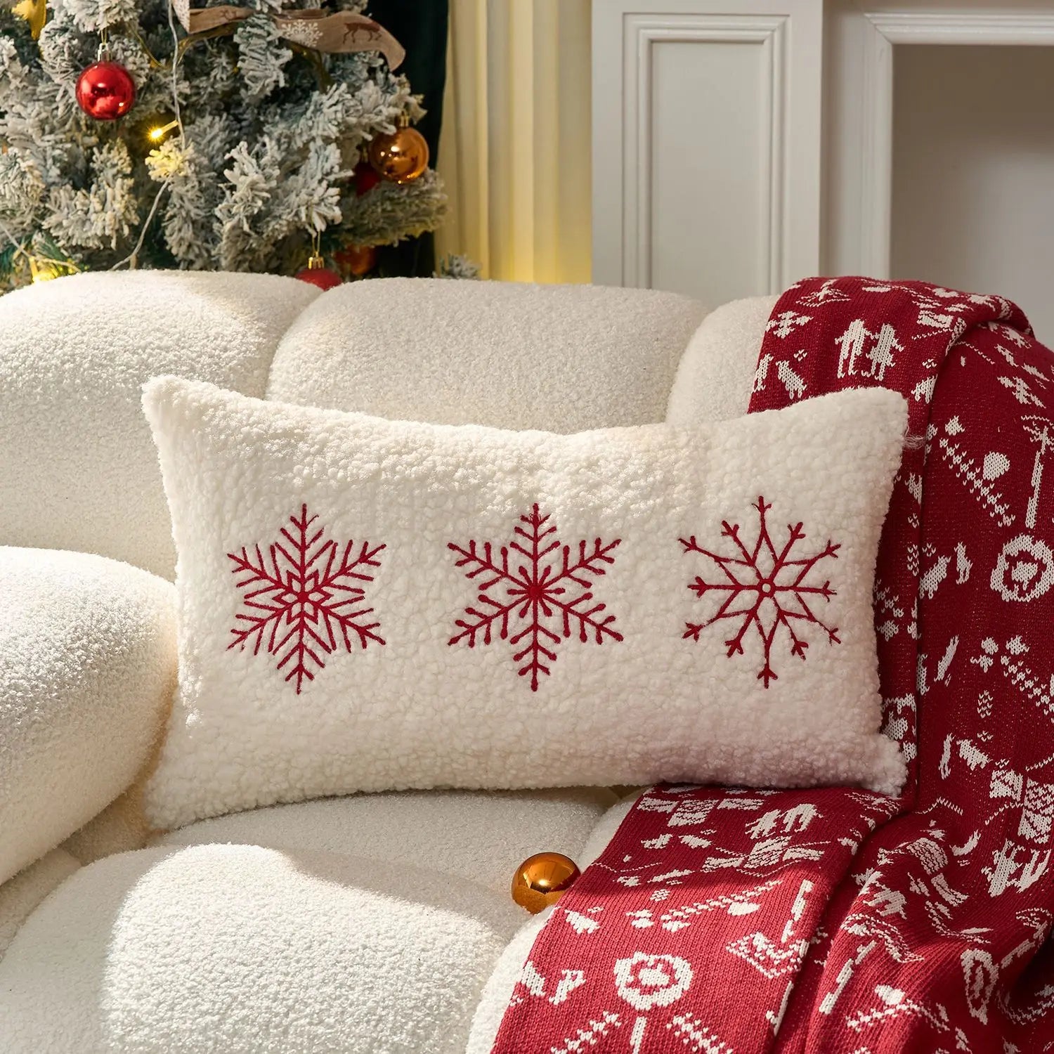 Decorative pillow with red snowflakes on a white couch with a red blanket and Christmas tree in the background.