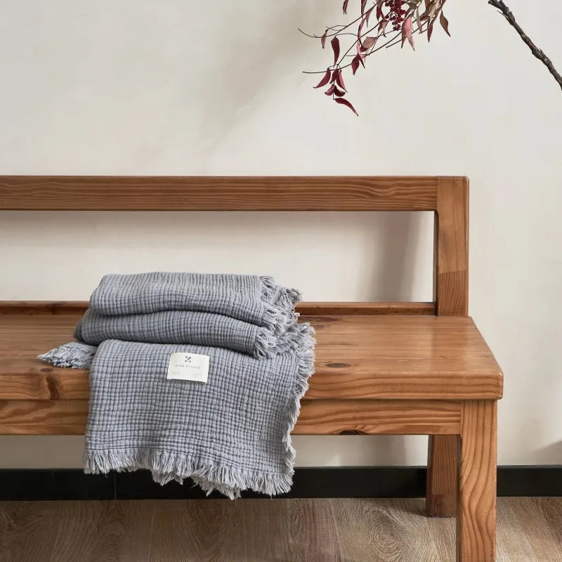 Wooden bench with a gray checkered blanket draped over it against a plain wall. Maillard cotton gauze blanket with tassels, four-layer muslin throw for sofa, bed, and boho home decor