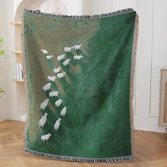 Green blanket with white sheep design on a wooden floor Grassland sheep tassel blanket, woven scenic throw for sofa, bed, or outdoor picnic, boho-inspired home decor blanket.