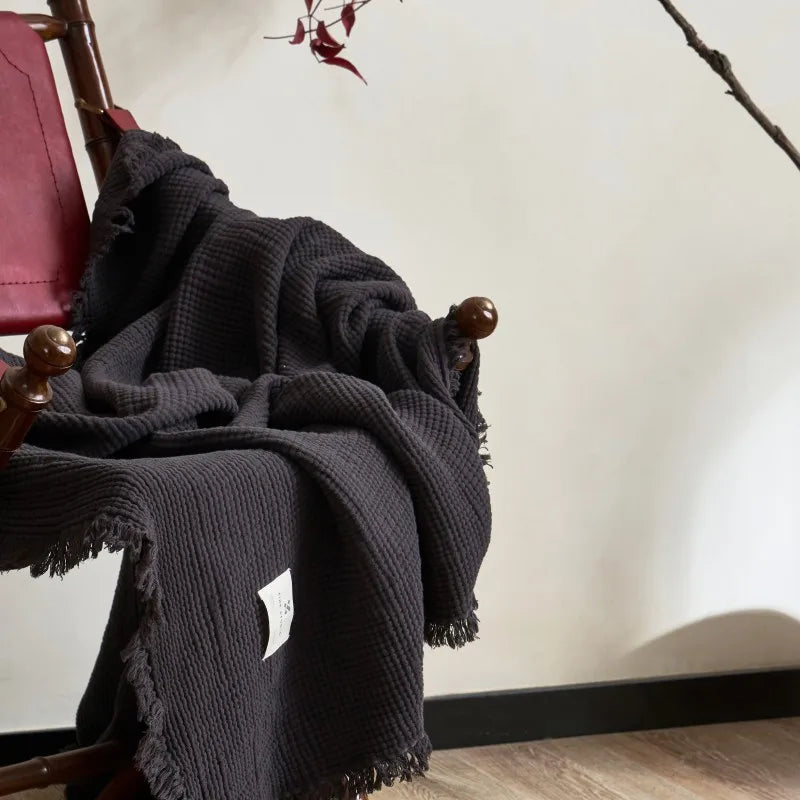 Black blanket draped over a wooden chair with a neutral background Maillard cotton gauze blanket with tassels, four-layer muslin throw for sofa, bed, and boho home decor