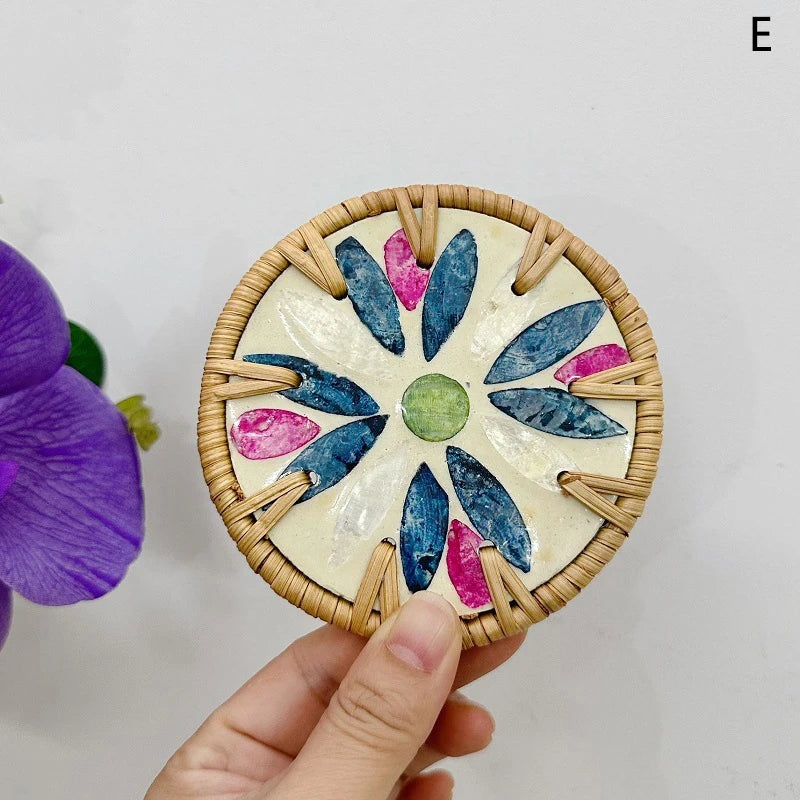 Decorative coaster with wicker rim and colorful stone design held by a hand, with a purple flower in the corner.
