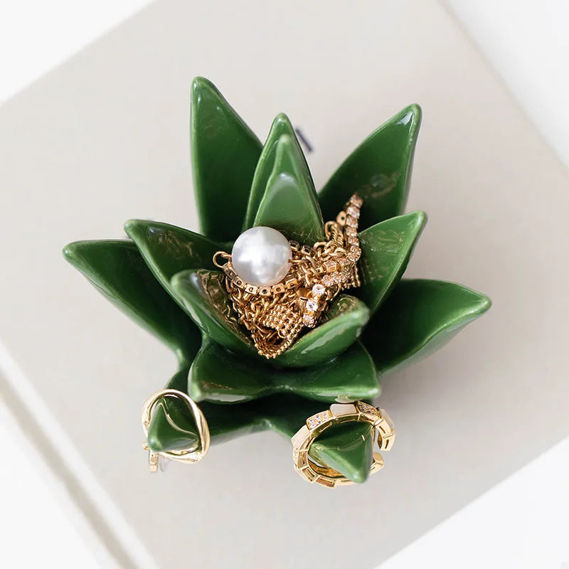 Green ceramic succulent-shaped jewelry holder with gold and pearl earrings on a white surface. Green ceramic cactus jewelry storage dish for home decor