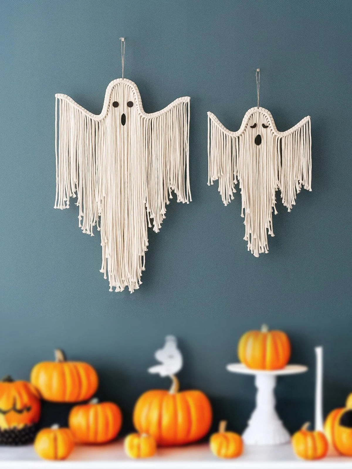 Two cream macrame ghost wall hangings with fringe details, displayed on a dark teal wall above a styled holiday table with pumpkins and a white bird figurine from My Boho Home