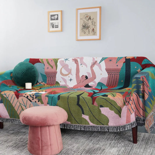 Colorful patterned sofa with a pink ottoman in a room with framed artwork on the wall. Boho Forest Dancer throw blanket with jungle leaves design and tassels
