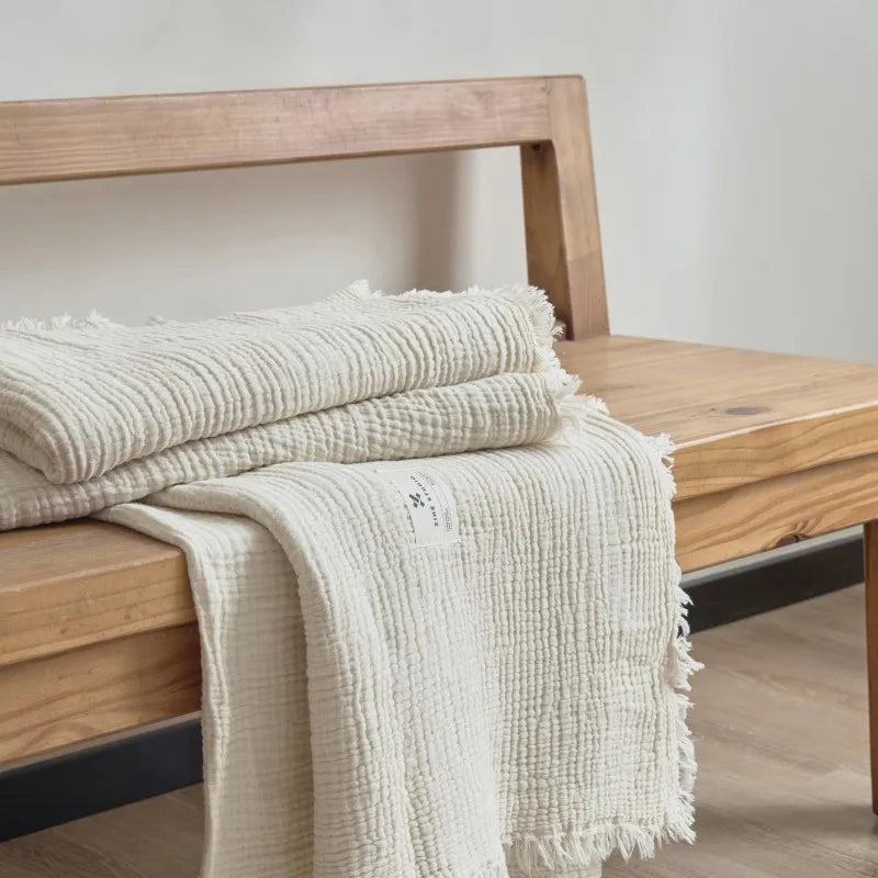 Stack of textured towels on a wooden bench with a neutral background Maillard cotton gauze blanket with tassels, four-layer muslin throw for sofa, bed, and boho home decor