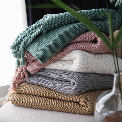 Stack of colorful boho knitted blankets on a surface with a plant in the foreground.