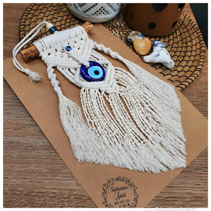 A handmade macrame wall hanging with long cream cords, a blue and white evil eye motif, and a wooden dowel, displayed on a brown kraft paper sheet over a woven mat.