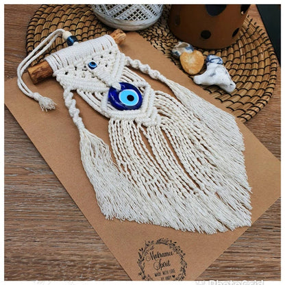 A handmade macrame wall hanging with long cream cords, a blue and white evil eye motif, and a wooden dowel, displayed on a brown kraft paper sheet over a woven mat.