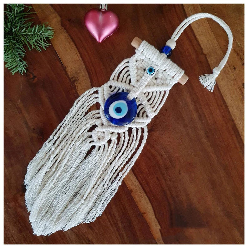 A white macrame wall hanging ornament shaped like a cat, featuring a large blue and white evil eye charm in the center, wooden dowel ends, and a hanging loop, displayed on a wooden surface.