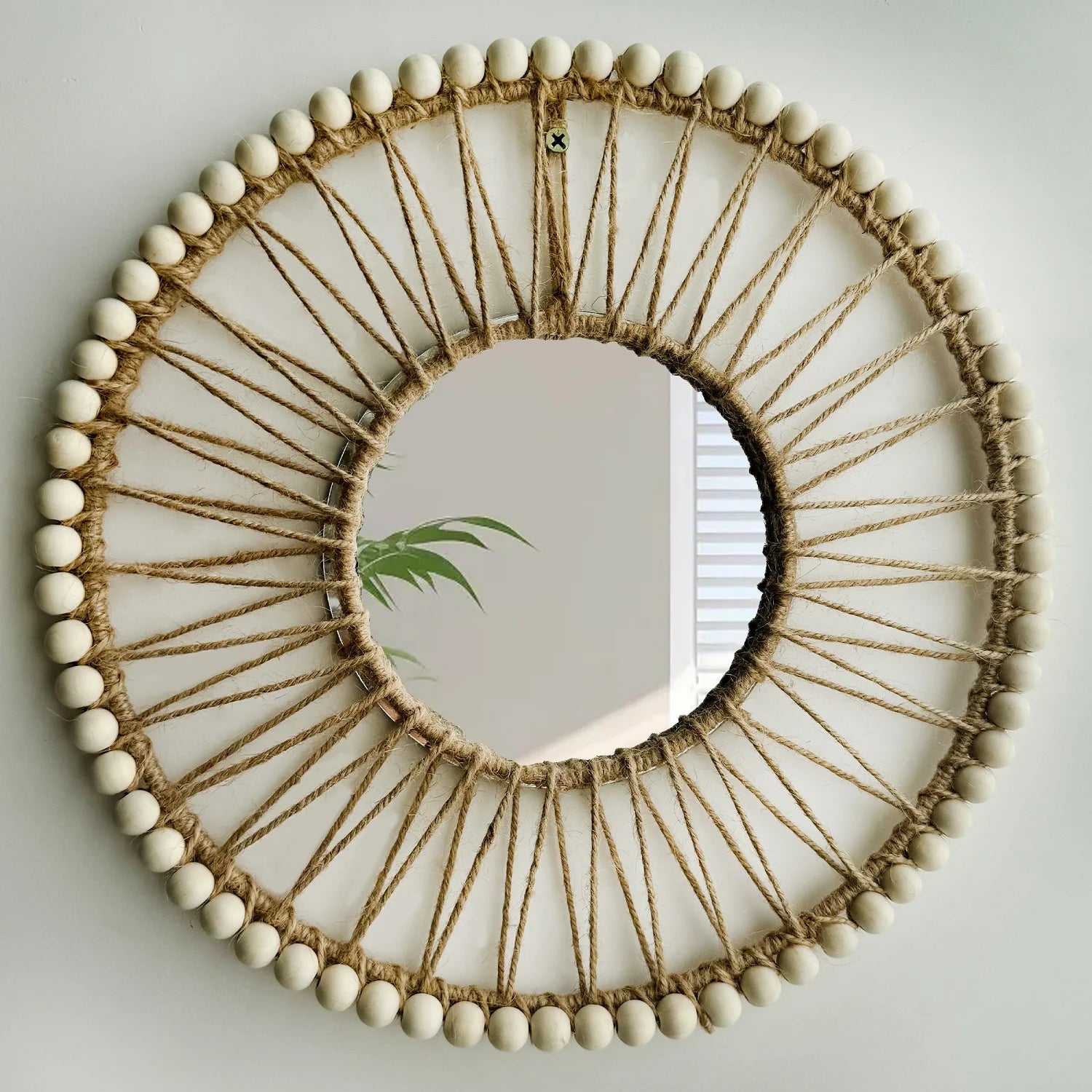 Round mirror with decorative frame made of twine and beads on a plain wall. Hand-woven Nordic round decorative mirror with Lafite grass frame and hemp rope, boho wall hanging for living room, entryway, or bedroom decor