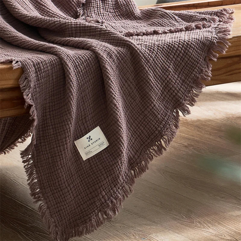 Brown textured blanket draped over a wooden surface with a visible label. Maillard cotton gauze blanket with tassels, four-layer muslin throw for sofa, bed, and boho home decor