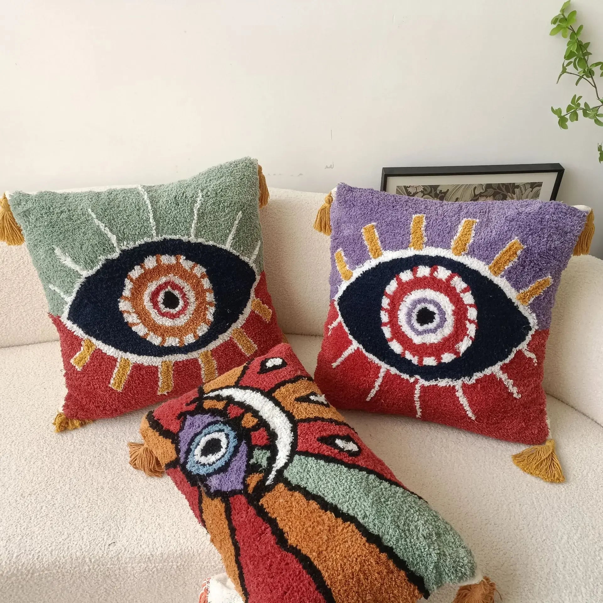 Three colorful decorative pillows with eye designs on a white couch. Boho Devil's Eye embroidered pillow cover with tufted geometric design in cotton blend fabric, decorative cushion for sofa, bed, or living room boho home decor.