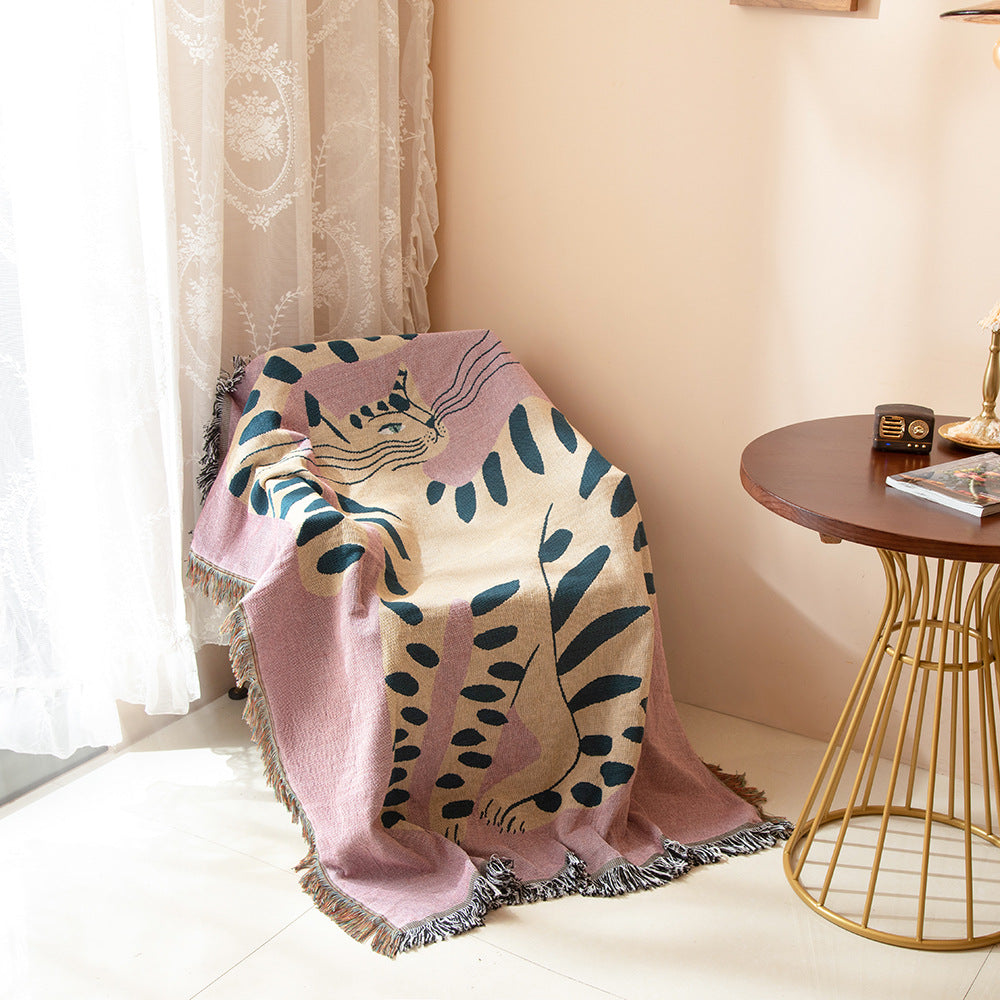 Decorative blanket with leaf pattern draped over a chair in a room with a side table and lamp. boho woven cotton tapestry with pink girl cat design