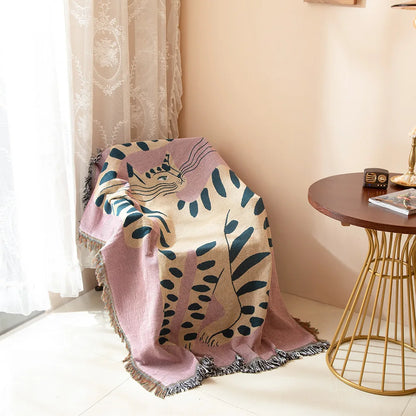 Decorative blanket with leaf pattern draped over a chair in a room with a side table and lamp. woven cotton tapestry with pink girl cat design