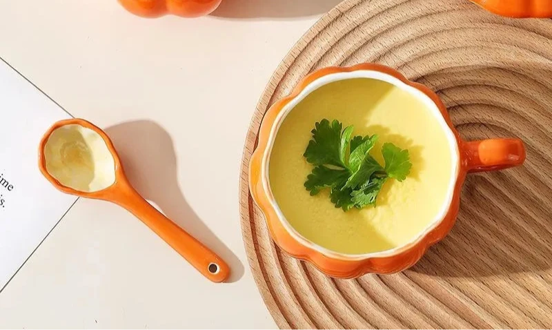 Orange pumpkin-shaped bowls with soup and a measuring spoon on a light background. Pumpkin ceramic mug with spoon Halloween autumn fall coffee cup