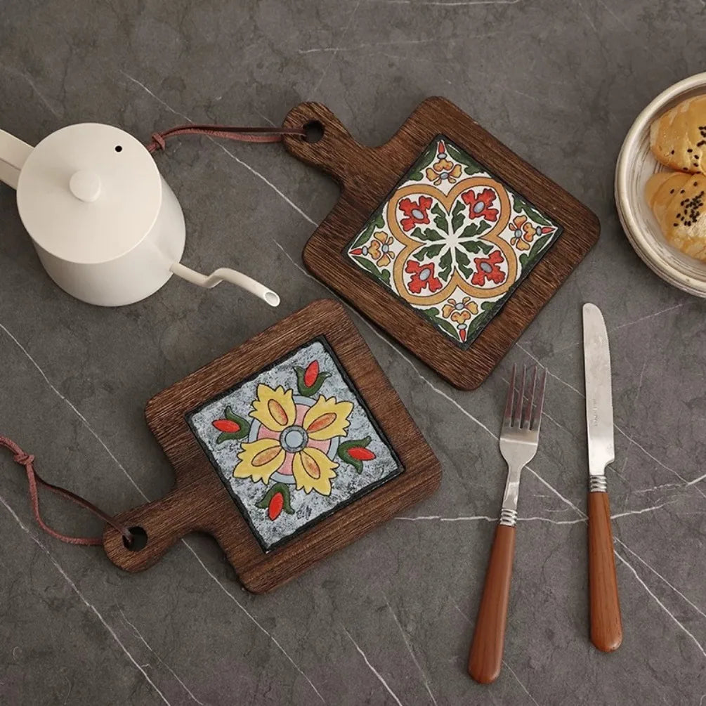Two wooden coasters with floral designs on a gray surface with a teapot and cutlery.