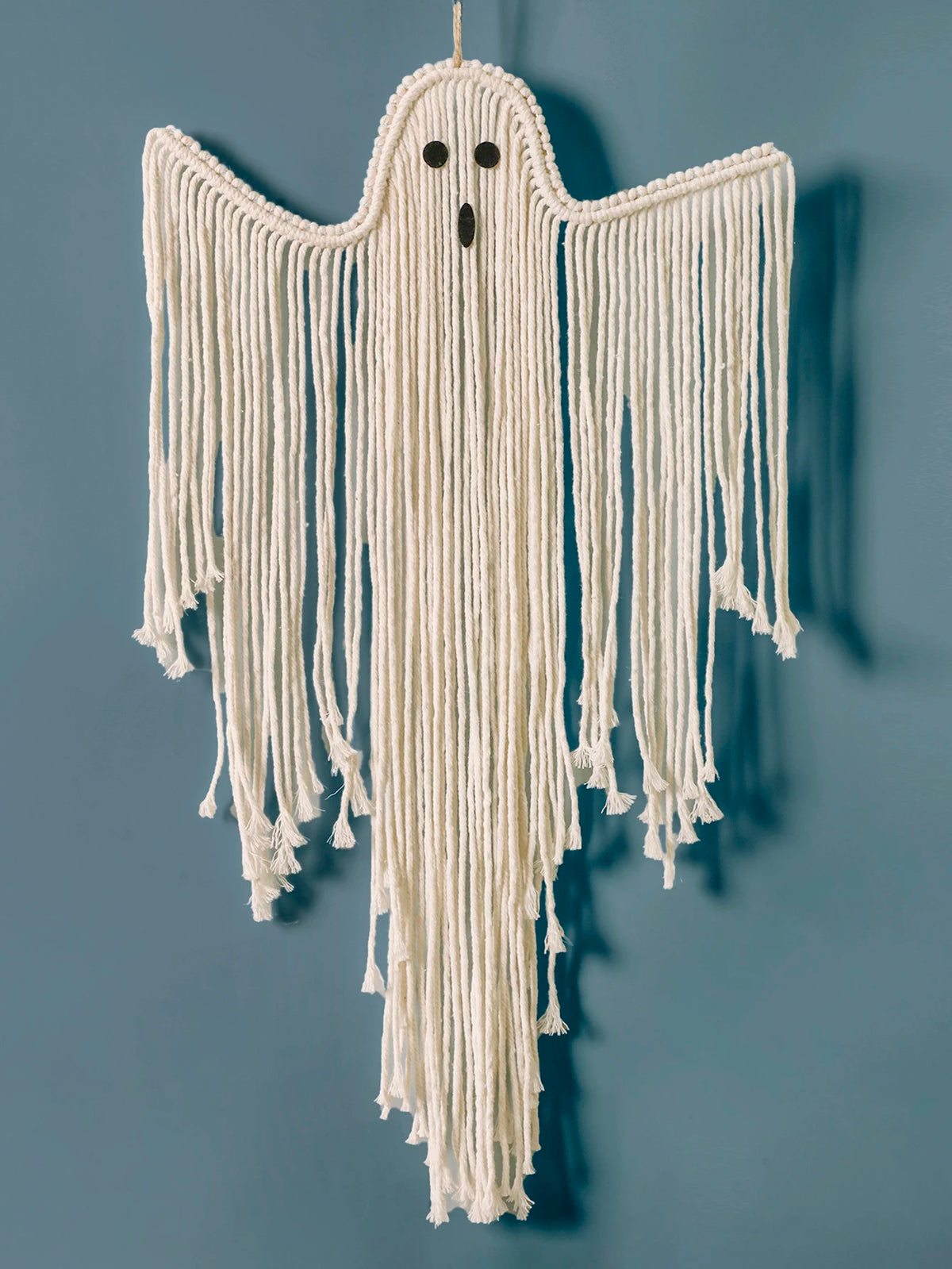 A ghost-shaped macrame wall hanging made from off-white yarn, featuring long fringe, small black embroidered eyes, and a black nose, shown hanging against a muted blue-gray wall from My Boho Home