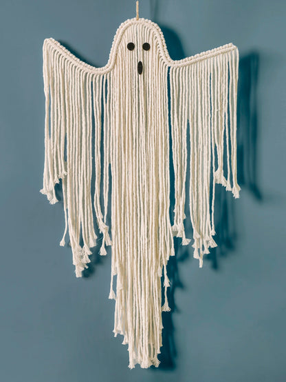 A ghost-shaped macrame wall hanging made from off-white yarn, featuring long fringe, small black embroidered eyes, and a black nose, shown hanging against a muted blue-gray wall from My Boho Home