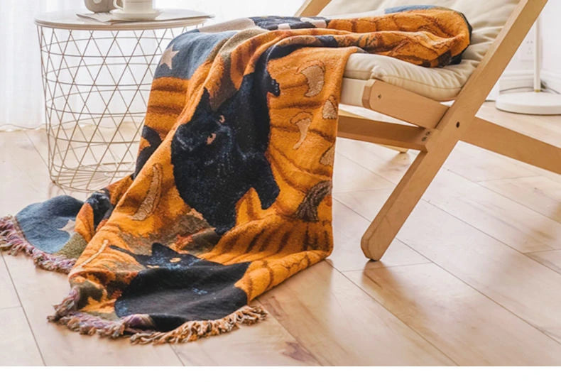 Blanket with cat design draped over a chair in a room. Fringed tapestry throw blanket with black cats and pumpkins, spooky bohemian Halloween home accessory