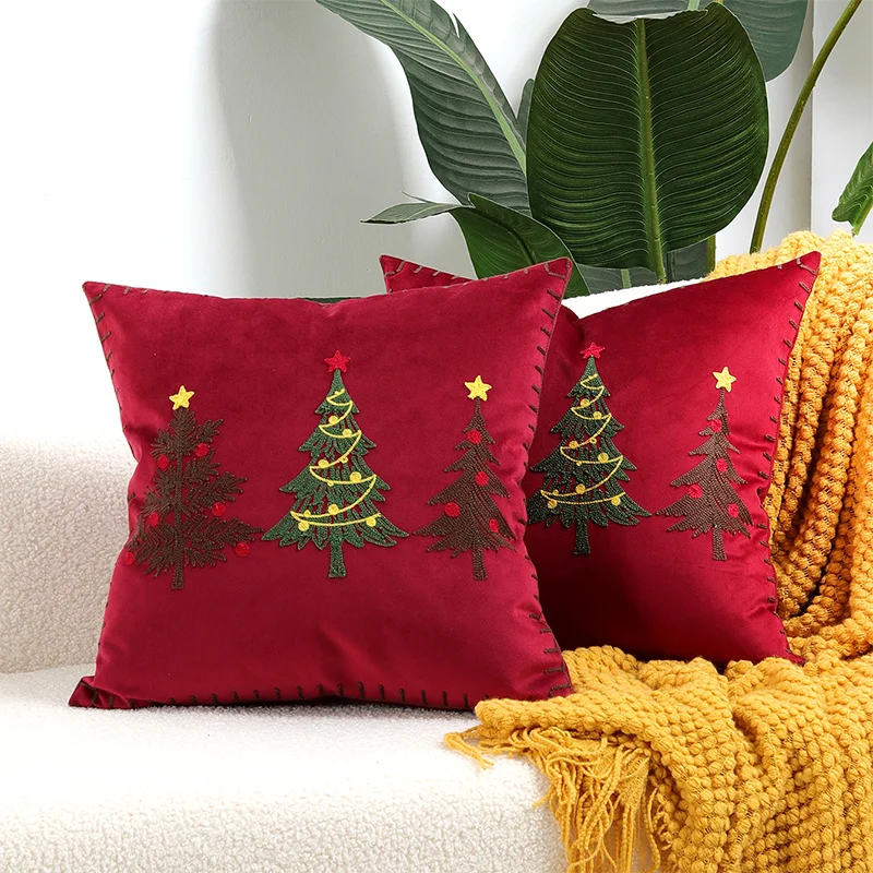Red pillows with Christmas tree designs on a couch with a yellow blanket and plant in the background. Embroidered Merry Christmas pillow cover with snowflake design – festive red and white holiday cushion for sofa or bedroom décor.
