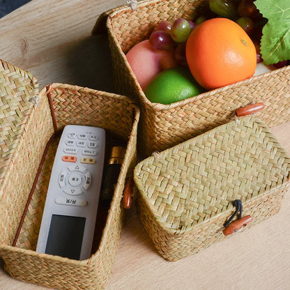 Woven baskets containing a remote control and fruits on a wooden surface Handmade seagrass rattan storage box with lid, rectangular woven organizer basket for makeup, jewelry, office, and boho home decor