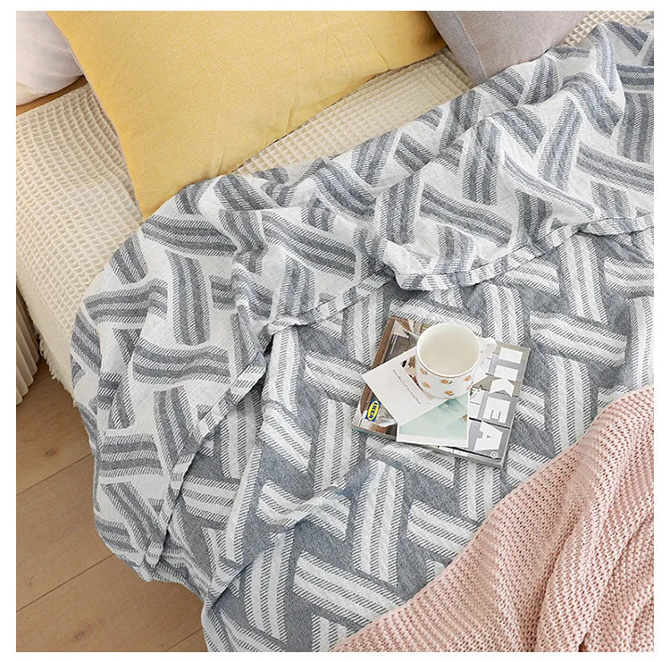 Gray and white chevron patterned blanket draped over a chair with a cup and magazine on it. Boho Lightweight microfiber summer blanket for bed or sofa, cooling breathable throw for home, dorm, or office naps with geometric jacquard pattern.