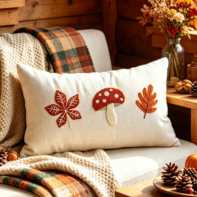 A square cream throw pillow with embroidered autumn motifs (a red maple leaf, a red-and-white mushroom, and an orange leaf) resting on a cozy beige couch in a rustic wood-paneled room with plaid blankets and fall decor. Boho fall home decor.