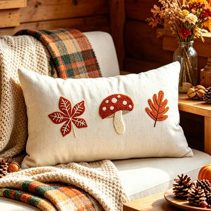 A square cream throw pillow with embroidered autumn motifs (a red maple leaf, a red-and-white mushroom, and an orange leaf) resting on a cozy beige couch in a rustic wood-paneled room with plaid blankets and fall decor. Boho fall home decor.