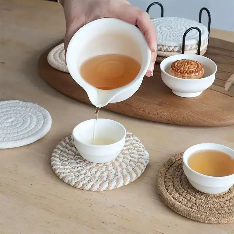 Person pouring tea from a white ceramic pot into a white ceramic cup on a wooden table with coasters. Hand placing a coffee mug on woven cotton coaster – practical and stylish table protection