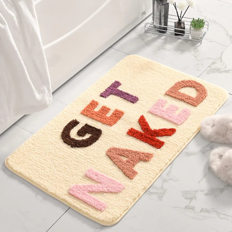 Beige bath mat with colorful letters spelling 'GET NAKED' on a bathroom floor. boho home decor, bathroom rug, tufted bath mat, Get Naked rug, colorful shower mat, apartment decor, dorm bathroom accessories, housewarming gift, microfiber bath mat, fun bathroom rug, American style bath mat