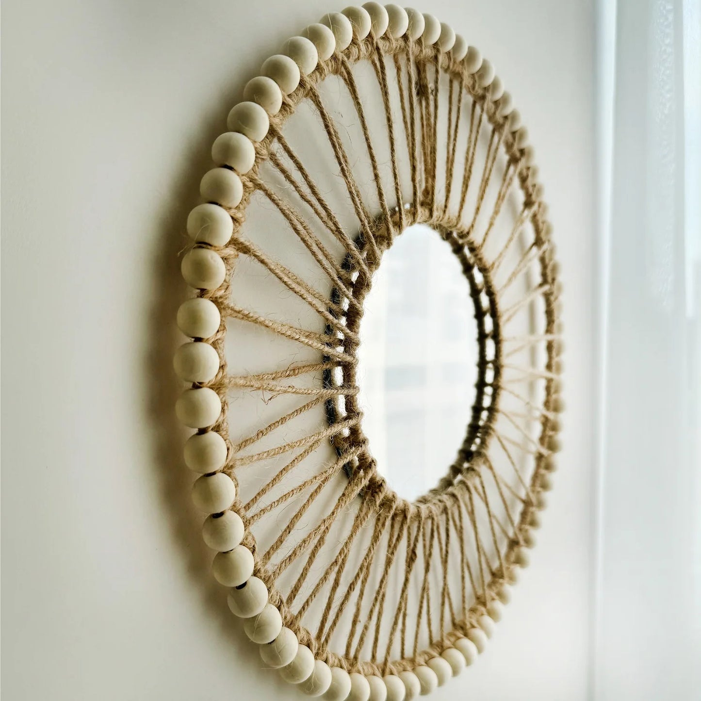 Decorative mirror with wooden beads and jute rope on a plain wall Hand-woven Nordic round decorative mirror with Lafite grass frame and hemp rope, boho wall hanging for living room, entryway, or bedroom decor