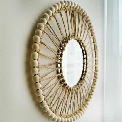 Decorative mirror with wooden beads and jute rope on a plain wall Hand-woven Nordic round decorative mirror with Lafite grass frame and hemp rope, boho wall hanging for living room, entryway, or bedroom decor